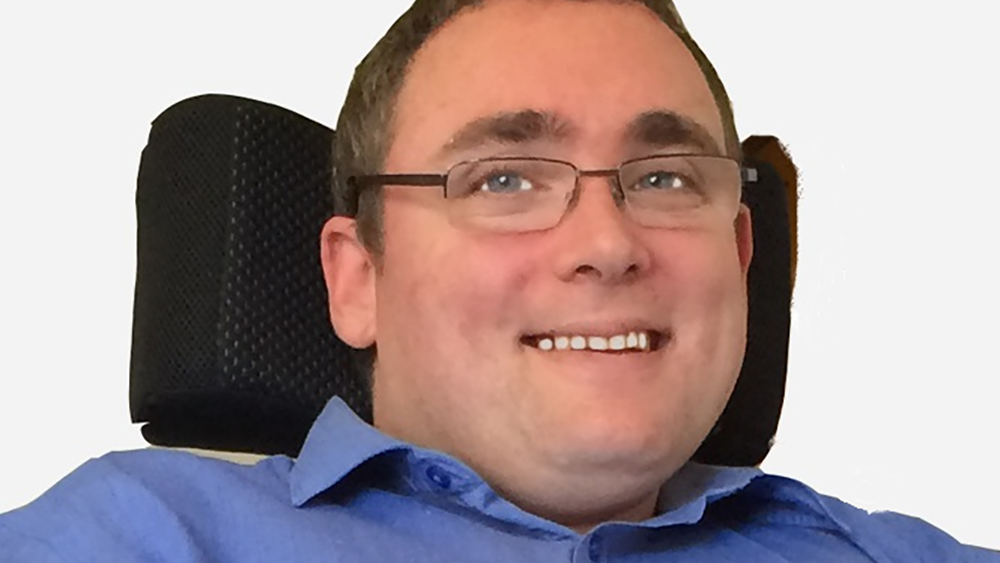 smiling headshot photo of ncat Board member Damian Bridgeman, wearing blue shirt and glasses sitting in a chair