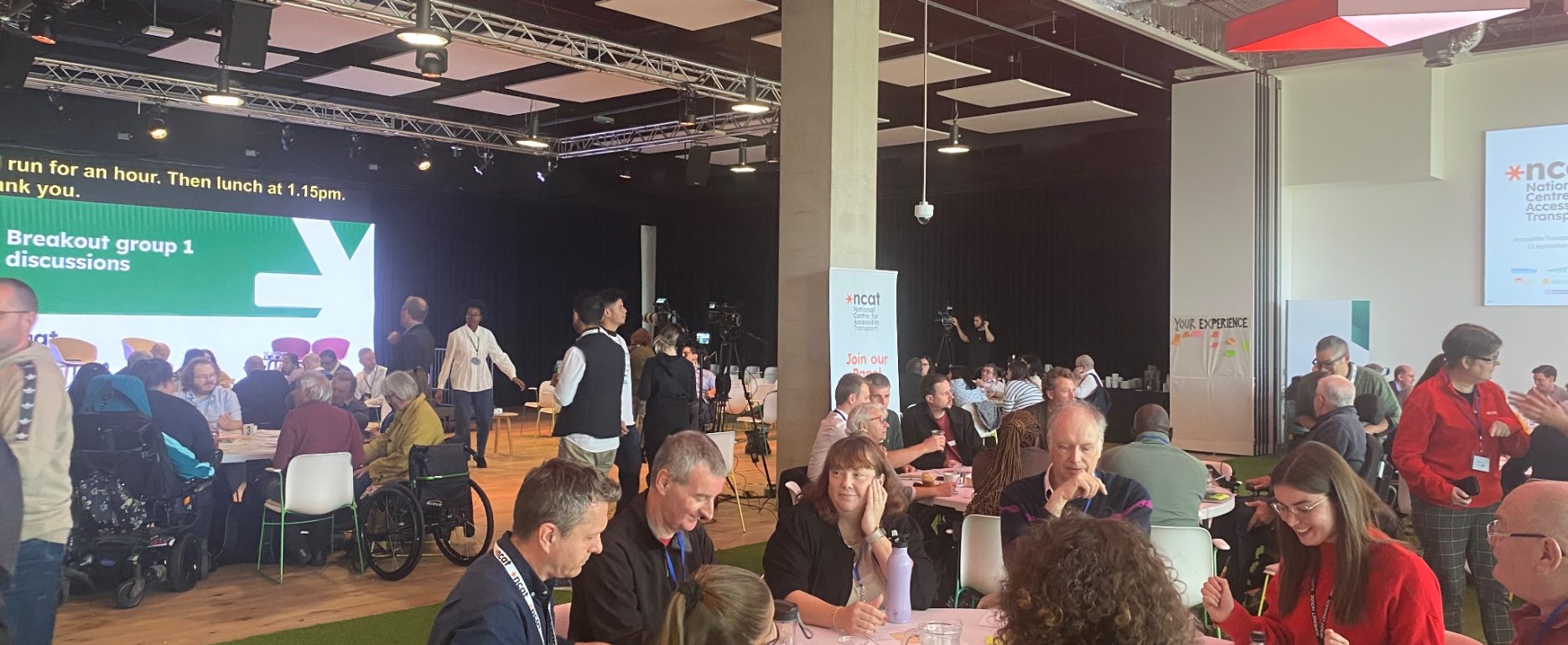 Large room with three large tables, around which there are between 8 to 10 participants sitting and discussing. In the background is a large screen reading 'Breakout group 1 discussions'.