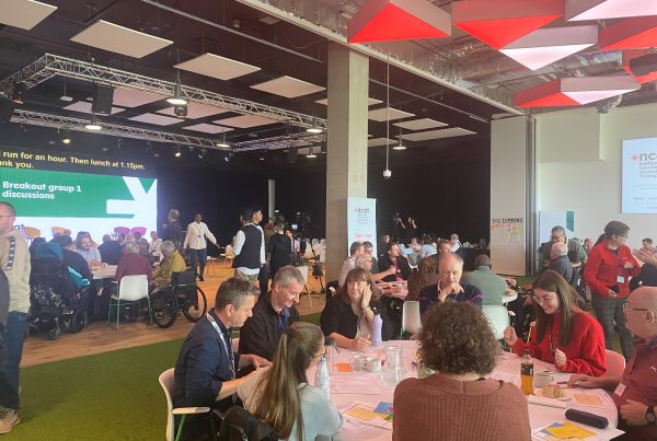 Large room with three large tables, around which there are between 8 to 10 participants sitting and discussing. In the background is a large screen reading 'Breakout group 1 discussions'.