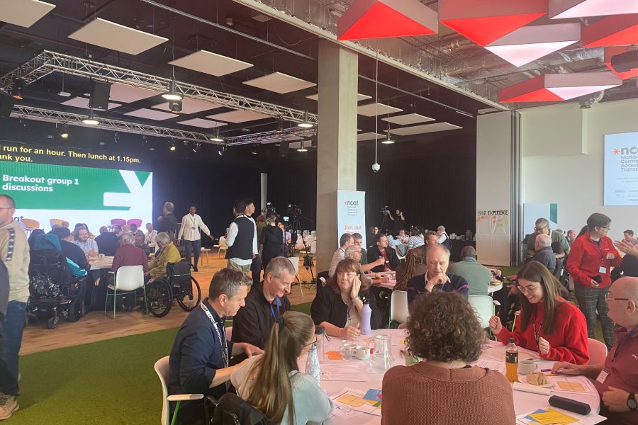 Large room with three large tables, around which there are between 8 to 10 participants sitting and discussing. In the background is a large screen reading 'Breakout group 1 discussions'.