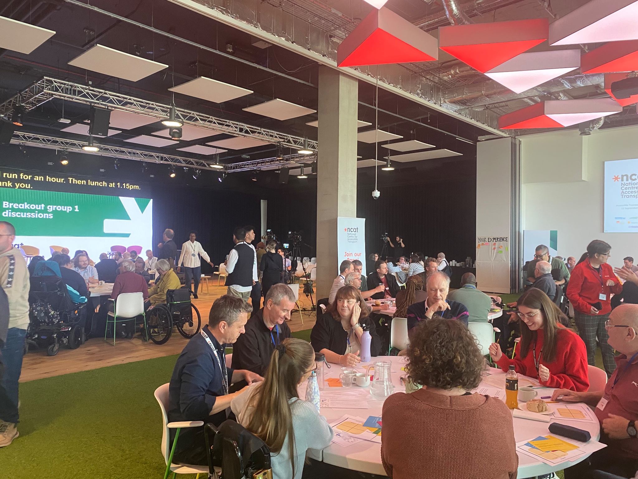 Large room with three large tables, around which there are between 8 to 10 participants sitting and discussing. In the background is a large screen reading 'Breakout group 1 discussions'.