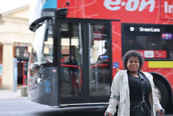 woman wearing coat and over the shoulder bag is standing facing the camera using mobility aids as support. Double decker bus is in background in front of brick building (Coventry Pool Meadow Bus Station.)