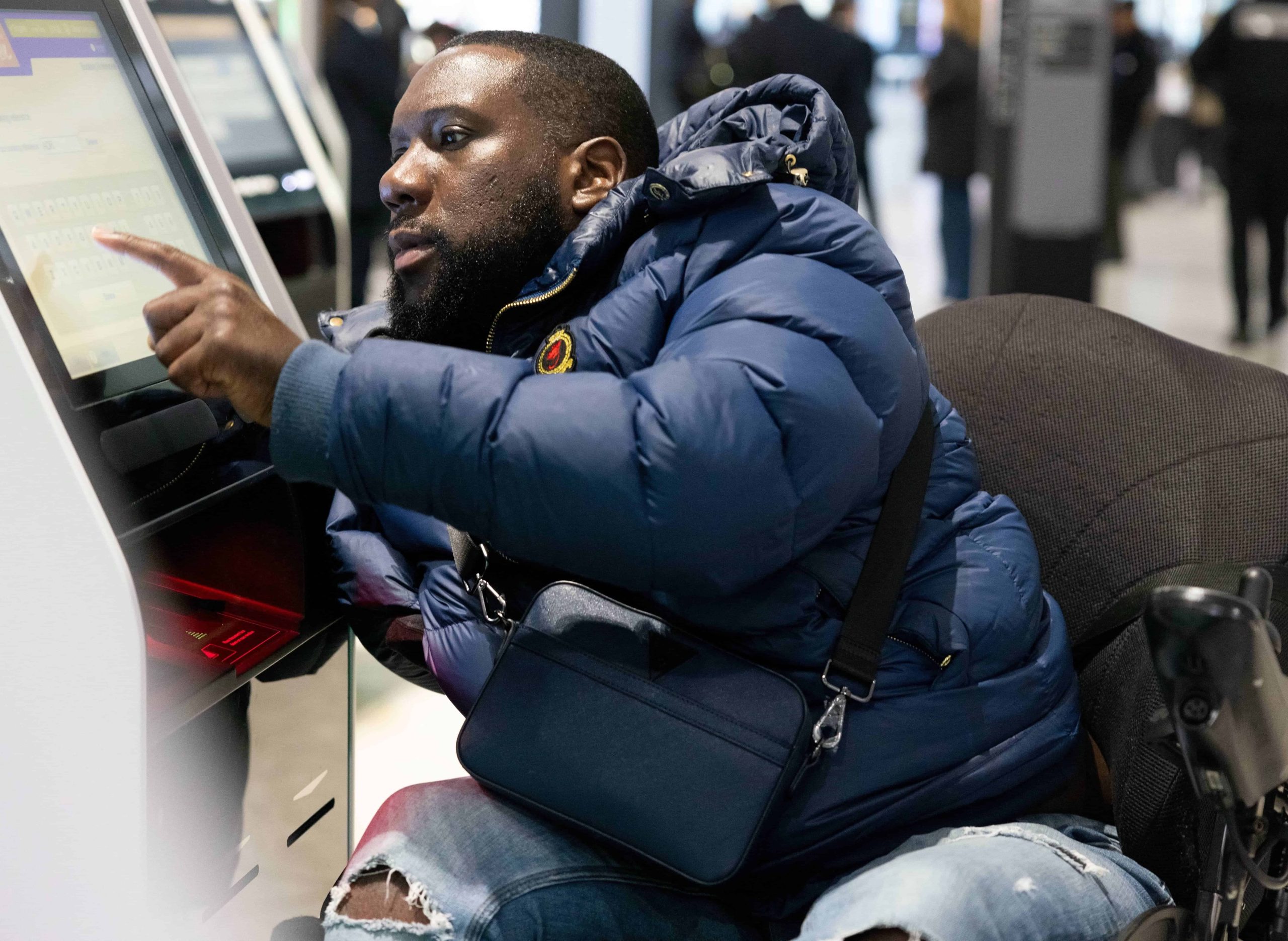 male wheelchair user wearing puffer jacket pressing screen on unit with left hand at Heathrow airport. About five people walking in the back of shot.