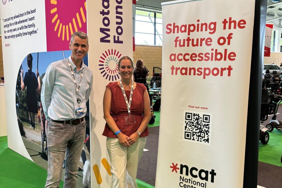 Paul Herriotts and Kay Atkin standing next to an ncat banner reading 'shaping the future of accessible transport'. A qr code is underneath the writing and above the ncat logo. This is in a large event space. Behind the banner there are some mobility vehicles and cars.