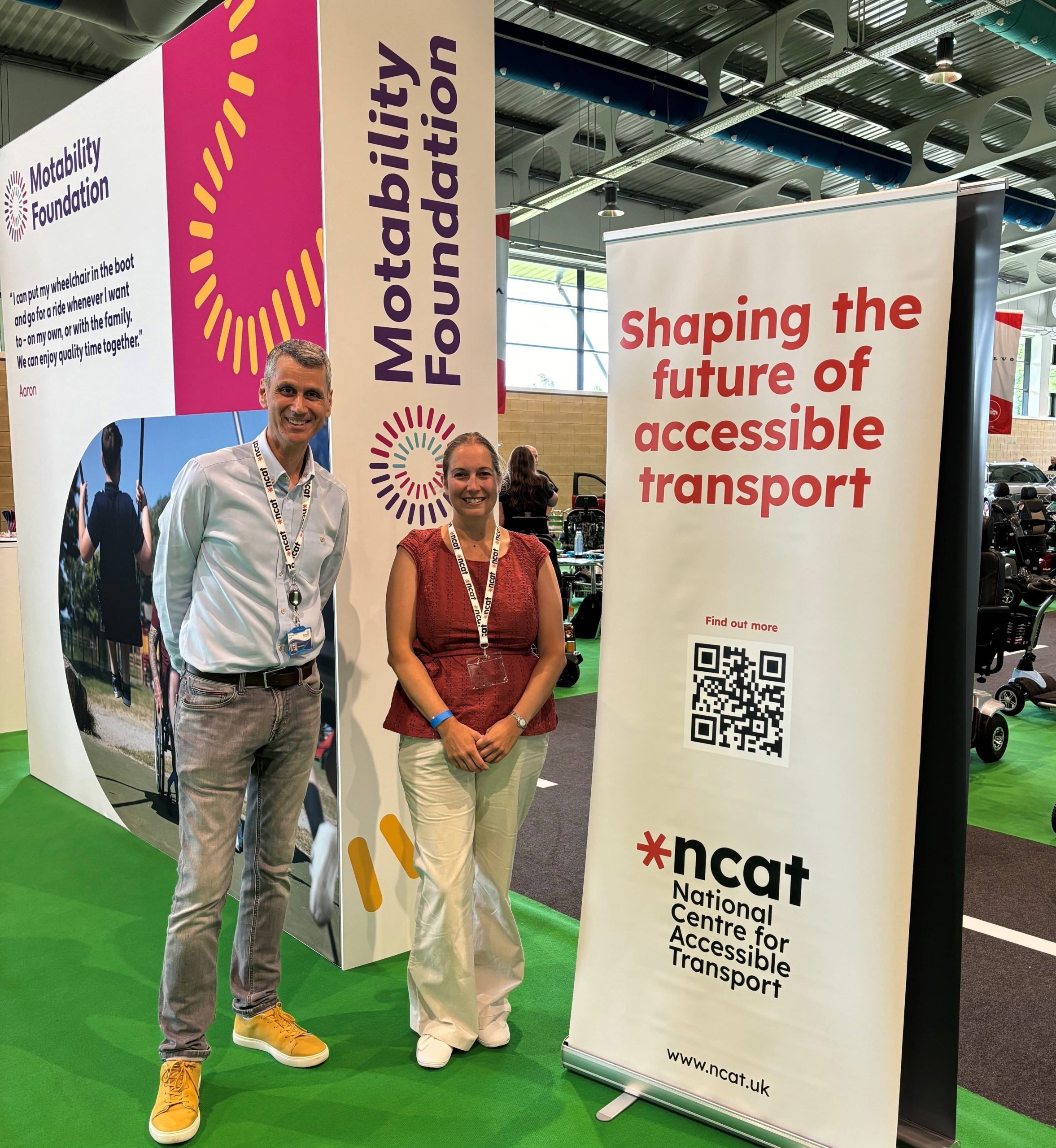 Paul Herriotts standing next to an ncat banner reading 'shaping the future of accessible transport'. A qr code is underneath the writing and above the ncat logo. This is in a large event space. Behind the banner there are some mobility vehicles and cars.