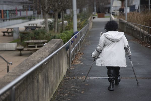 Woman in white coat walking down path using mobility aids. She has her back to the camera. There are handrails on either side of the path and on the left of the image are several picnic benches, bushes and trees.