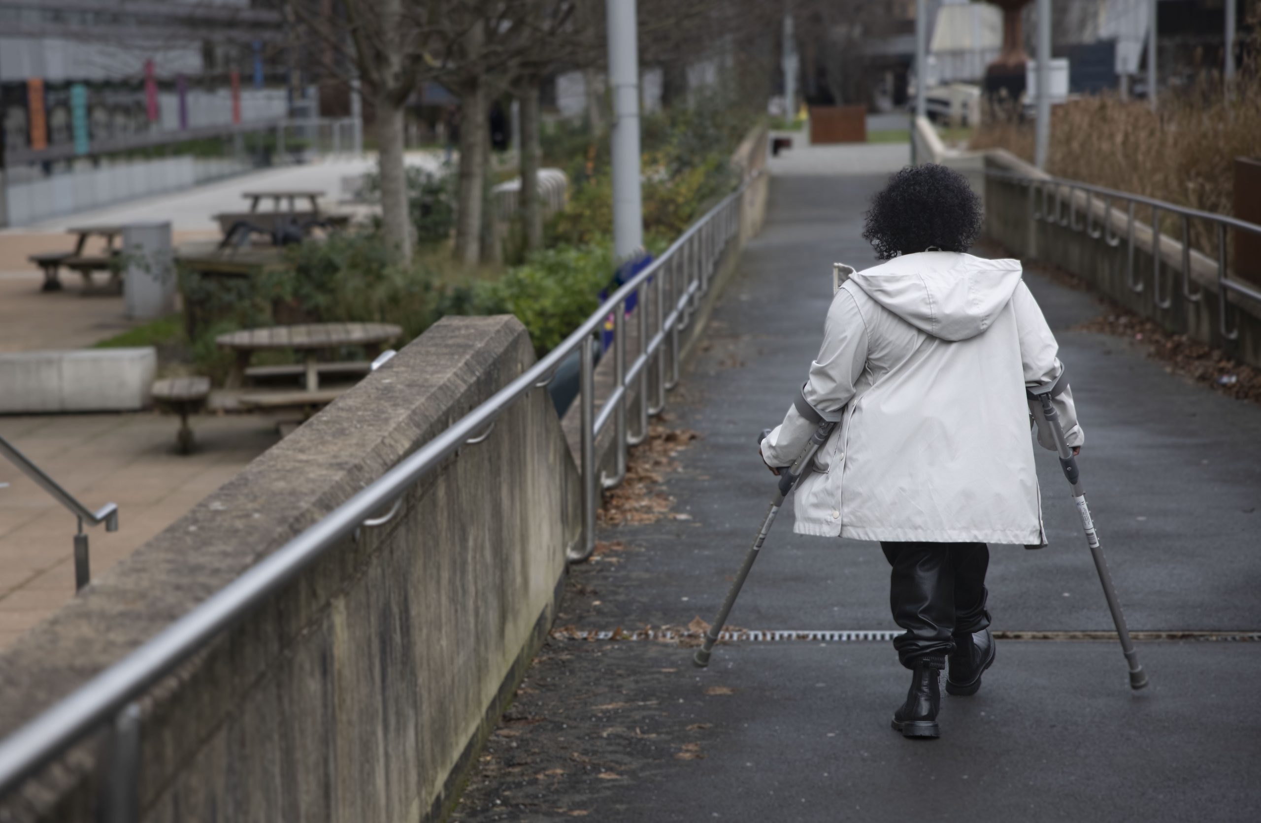 Woman in white coat walking down path using mobility aids. She has her back to the camera. There are handrails on either side of the path and on the left of the image are several picnic benches, bushes and trees.