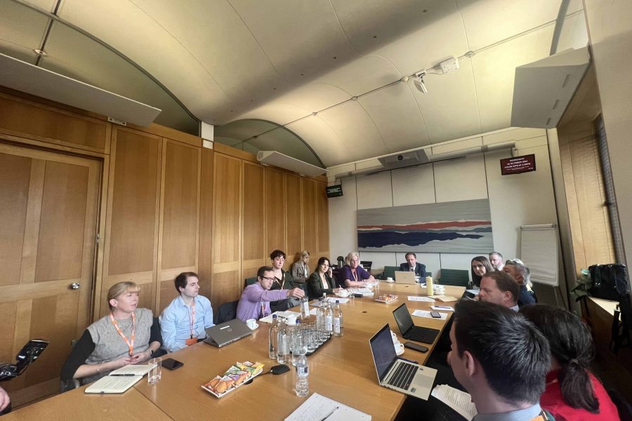 Long rectangular table down the middle of the image with about seven people sitting on one side, one at the head of the table, and about seven sitting on the right. There are open laptops and notebooks and water glasses on the table. The wall behind is wood panelled and there is a large abstract picture on the wall in the background.