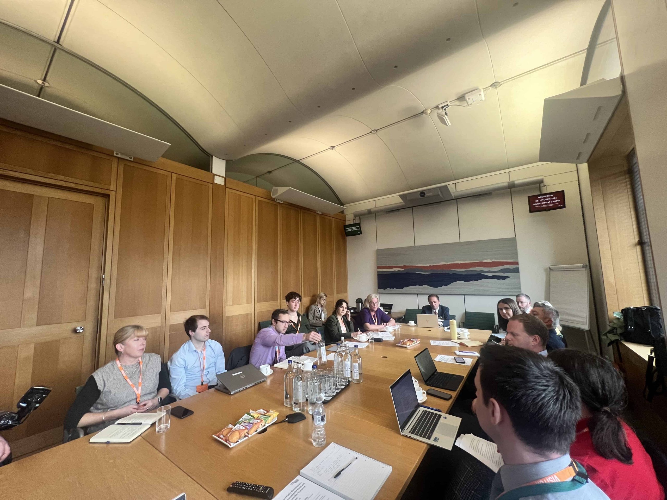 Long rectangular table down the middle of the image with about seven people sitting on one side, one at the head of the table, and about seven sitting on the right. There are open laptops and notebooks and water glasses on the table. The wall behind is wood panelled and there is a large abstract picture on the wall in the background.