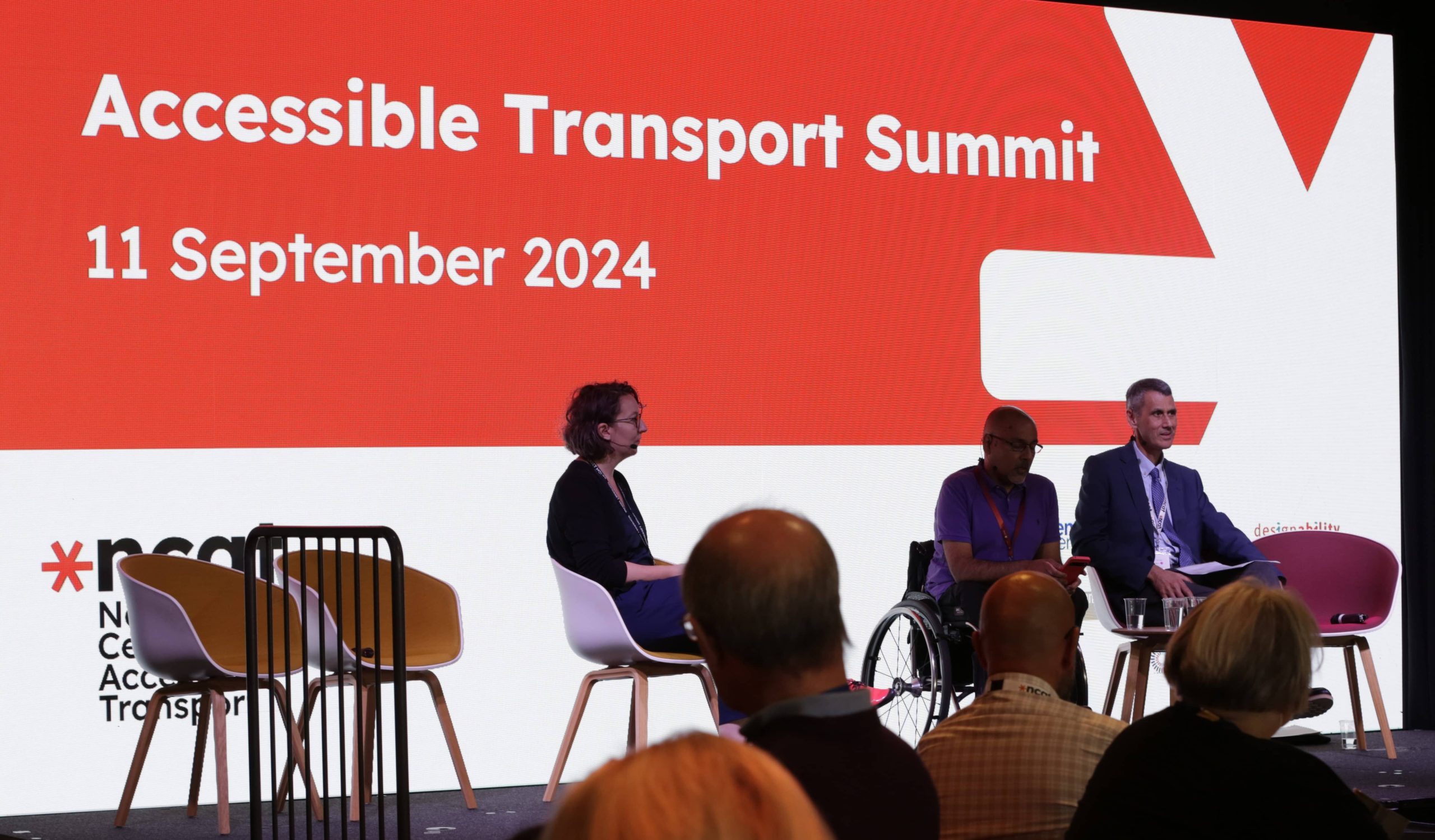 Lucy Farrow from Thinks Insight, Kamran Mallick from Disability Rights UK and Paul Herriotts from the National Centre for Accessible Transport are sitting on the stage in front of a large screen reading 'Accessible Transport Summit 11th September 2024'. There are three empty chairs also on the stage. In the foreground of the image we can see the backs of the heads of about four audience members as they are looking at the stage.