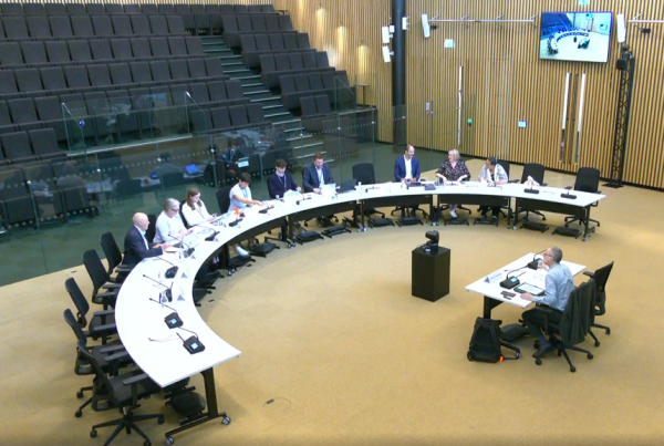 Tables arranged in a large semi circle with 18 chairs behind one side. Half the chairs are empty but nine are filled with a mix of men and women wearing office clothing. They are facing one small single table behind which one man is sitting. Behind all the tables are empty seats in lecture hall style.