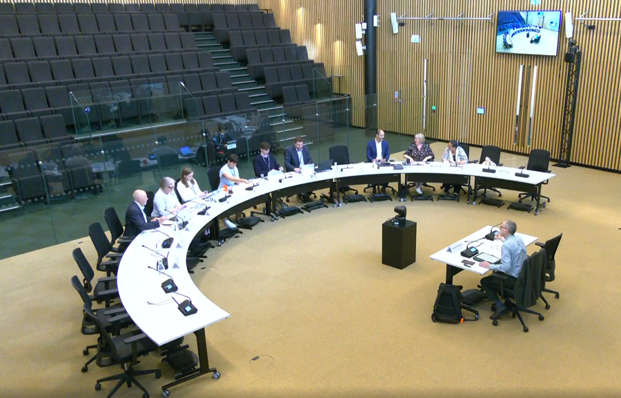 Tables arranged in a large semi circle with 18 chairs behind one side. Half the chairs are empty but nine are filled with a mix of men and women wearing office clothing. They are facing one small single table behind which one man is sitting. Behind all the tables are empty seats in lecture hall style.