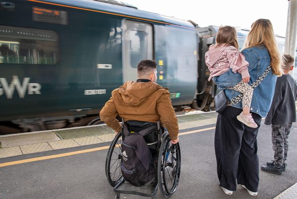 Family with their backs to the camera, waiting on a railway platform with train in front of them. One adult in a wheelchair, one adult holding a small cild, with another child alongside.