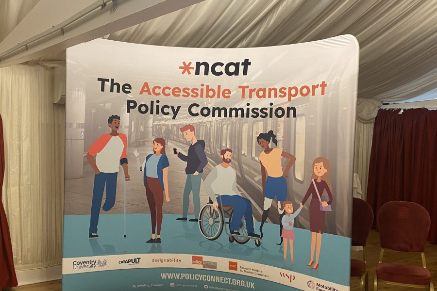 Standing ncat banner with 'The Accessible Transport Policy Commission' written across the top. Underneath are illustrations of six people representing diverse ethnicities and disabilities and genders. The logos of the consortium are written across the bottom of the banner. In the background are a couple of empty chairs and red curtains.