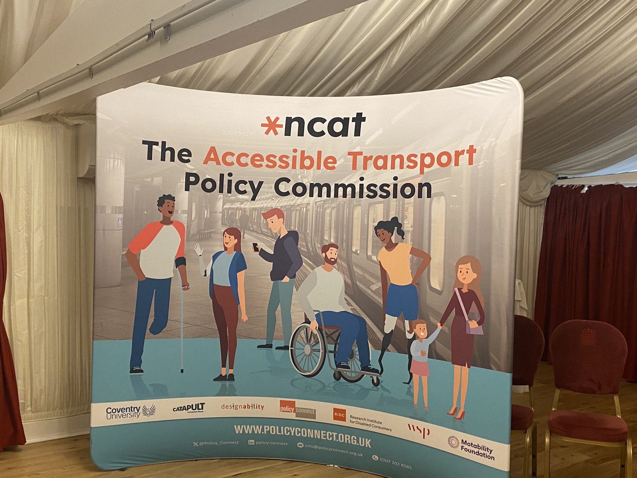 Standing ncat banner with 'The Accessible Transport Policy Commission' written across the top. Underneath are illustrations of six people representing diverse ethnicities and disabilities and genders. The logos of the consortium are written across the bottom of the banner. In the background are a couple of empty chairs and red curtains.