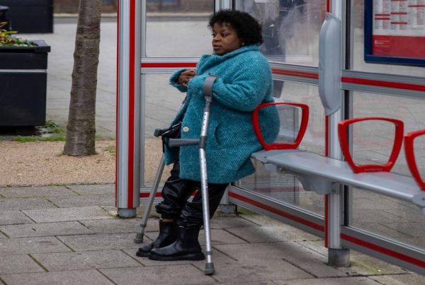 woman wearing blue coat leans against metal bench in sheltered bus stop while using mobility aids. A tree and bin are in the background of the image.