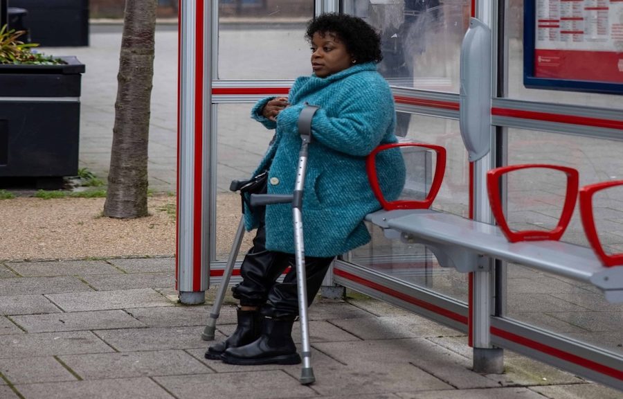 woman wearing blue coat leans against metal bench in sheltered bus stop while using mobility aids. A tree and bin are in the background of the image.