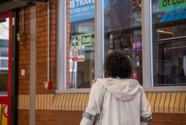 Woman in white coat walking using mobility aids in bus station. Double decker bus can be seen in background through windows. Screen in top left of image for bus times