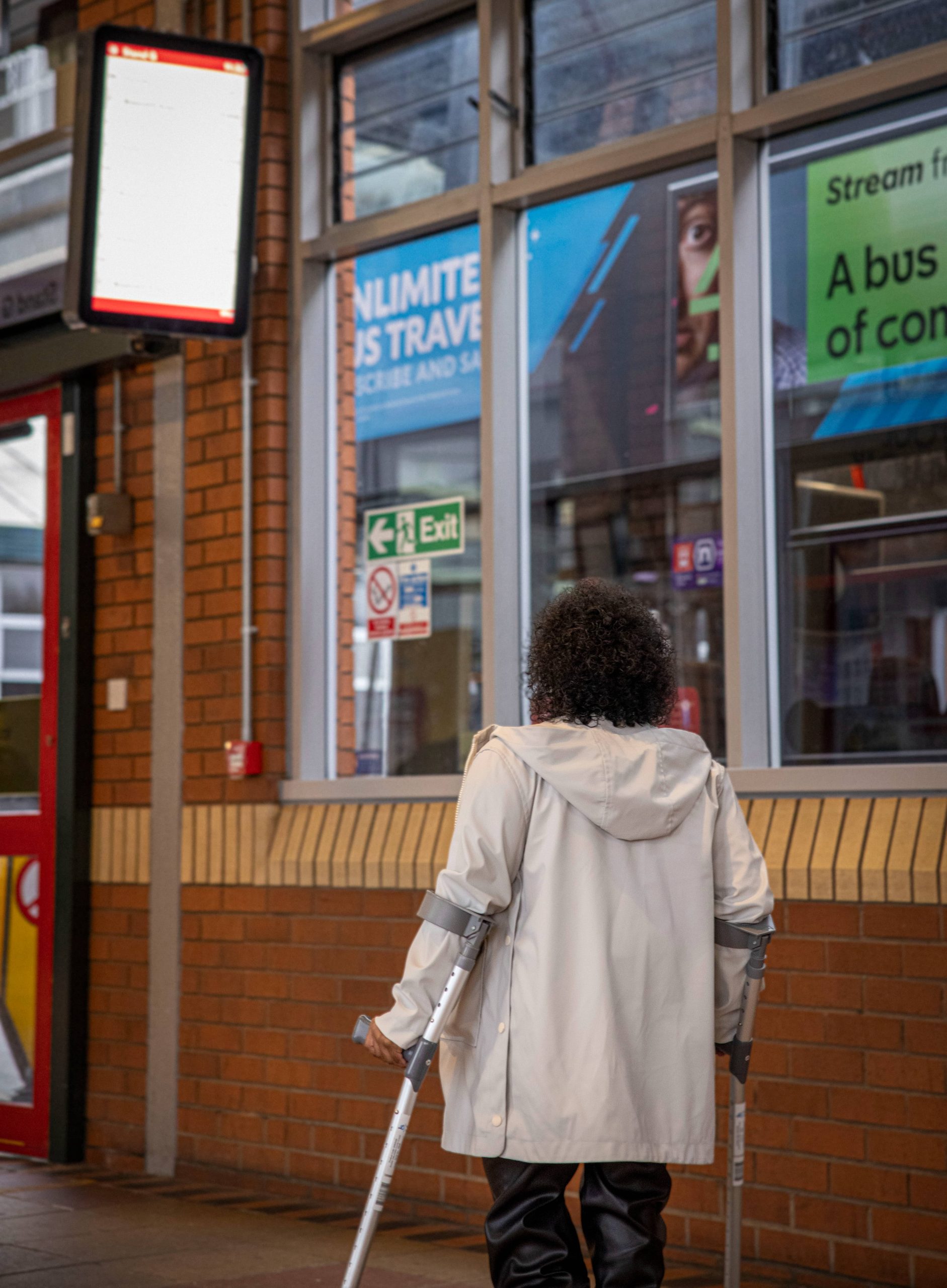 Woman in white coat walking using mobility aids in bus station. Double decker bus can be seen in background through windows. Screen in top left of image for bus times