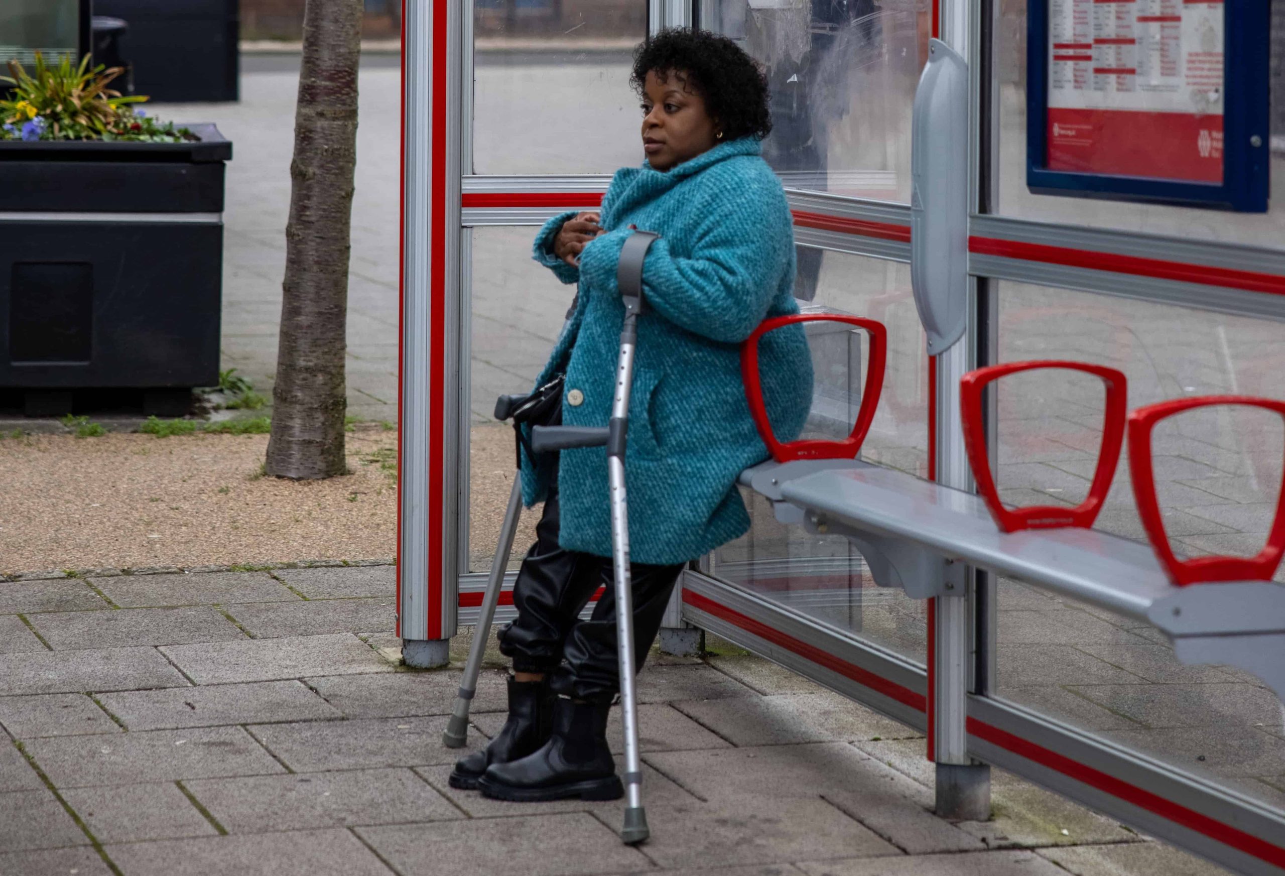 woman wearing blue coat leans against metal bench in sheltered bus stop while using mobility aids. A tree and bin are in the background of the image.